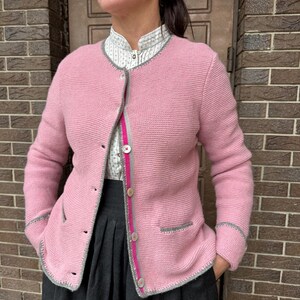Puede incluir: Un cárdigan rosa claro con ribetes y botones grises. El cárdigan tiene un cuello redondo y dos bolsillos delanteros. La modelo lleva una camisa blanca con cuello con un estampado gris y blanco y una falda gris oscuro.