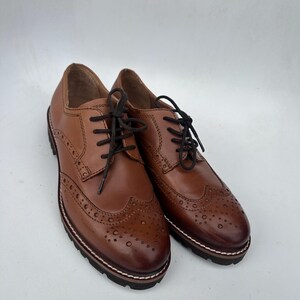 May include: A pair of brown leather brogue shoes with black laces. The shoes have decorative perforations and a thick, dark sole with white stitching. The shoes are displayed on a white background.
