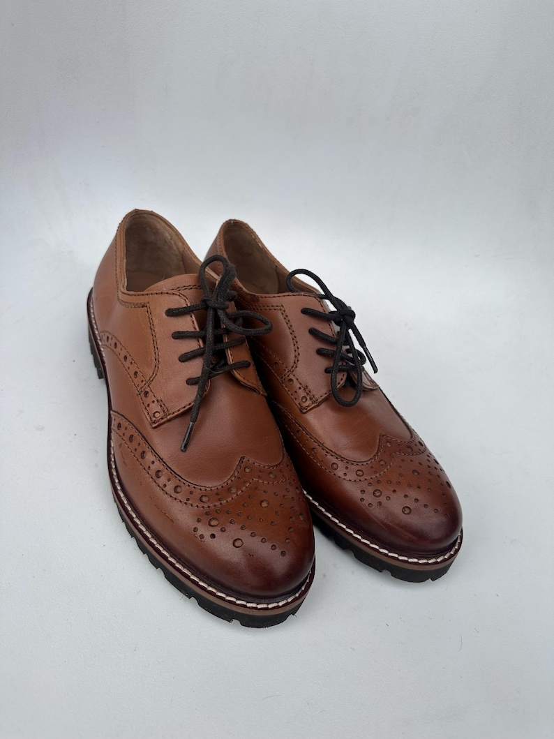 May include: A pair of brown leather brogue shoes with black laces. The shoes have decorative perforations and a thick, dark sole with white stitching. The shoes are displayed on a white background.