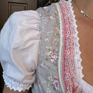 May include: A bodice featuring a grey fabric with a pink and green floral pattern, paired with white puff sleeves. The neckline and front panel are decorated with pink fabric trim, intricate white lace edging, and a scalloped white lace border.
