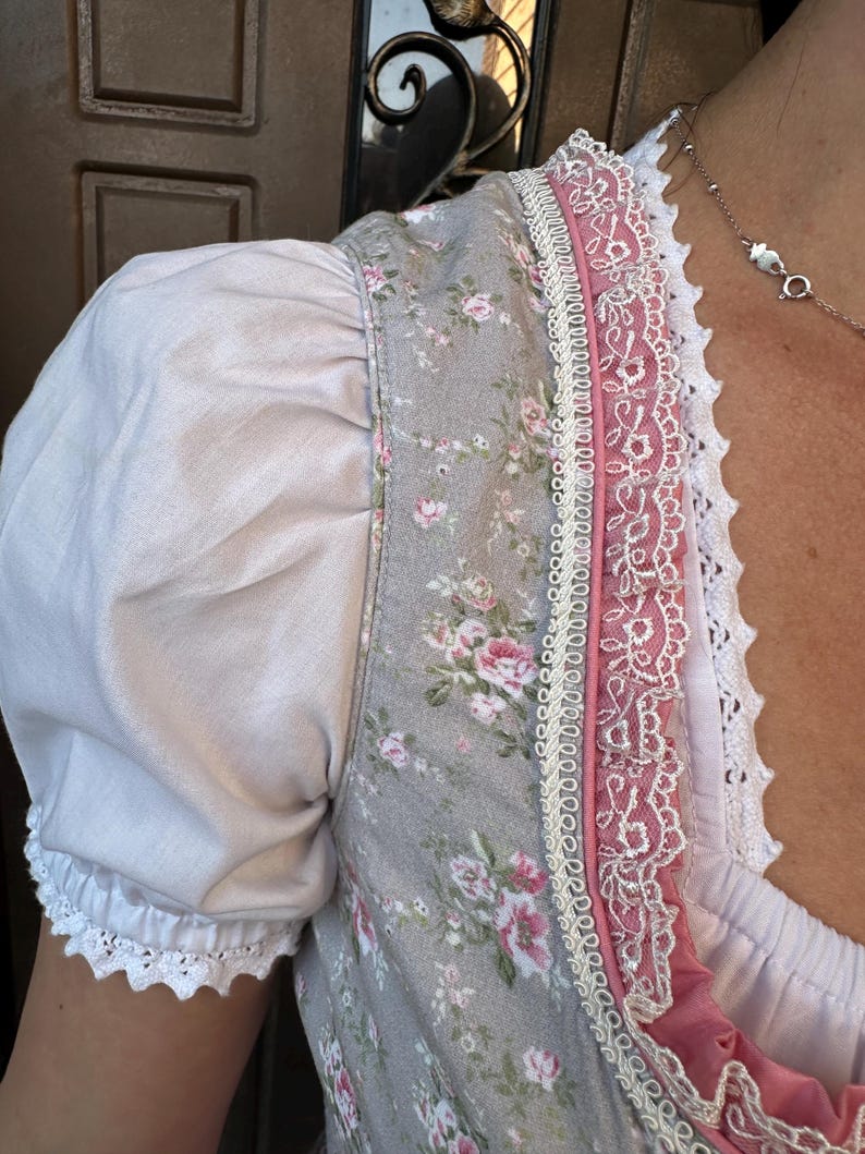 May include: A bodice featuring a grey fabric with a pink and green floral pattern, paired with white puff sleeves. The neckline and front panel are decorated with pink fabric trim, intricate white lace edging, and a scalloped white lace border.
