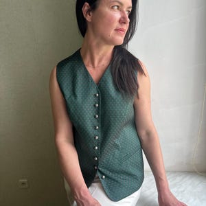 May include: A sleeveless green vest with a diamond pattern and silver buttons. The vest has a V-neck and a tailored fit. The fabric appears to be a woven material. The vest is worn over white pants.