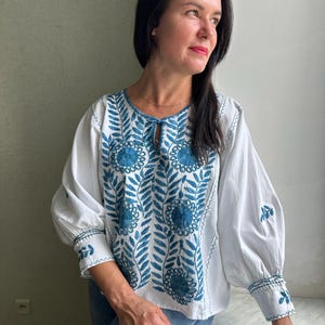 May include: A white blouse with blue embroidered floral designs. The blouse has a round neckline and long sleeves.
