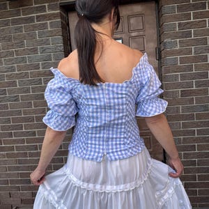Puede incluir: Un top azul y blanco de hombros descubiertos con estampado de cuadros vichy, peque&ntilde;as flores y mangas abullonadas, combinado con una falda blanca de volantes con encaje y cintas. El top tiene una cremallera en la parte trasera.