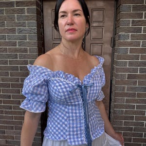 Puede incluir: Una blusa de hombros descubiertos en cuadros vichy azul claro y blanco, con mangas cortas abullonadas, escote coraz&oacute;n y un detalle de cordones con cinta azul en el centro. La tela tiene un estampado floral sutil y bordes de encaje blanco.