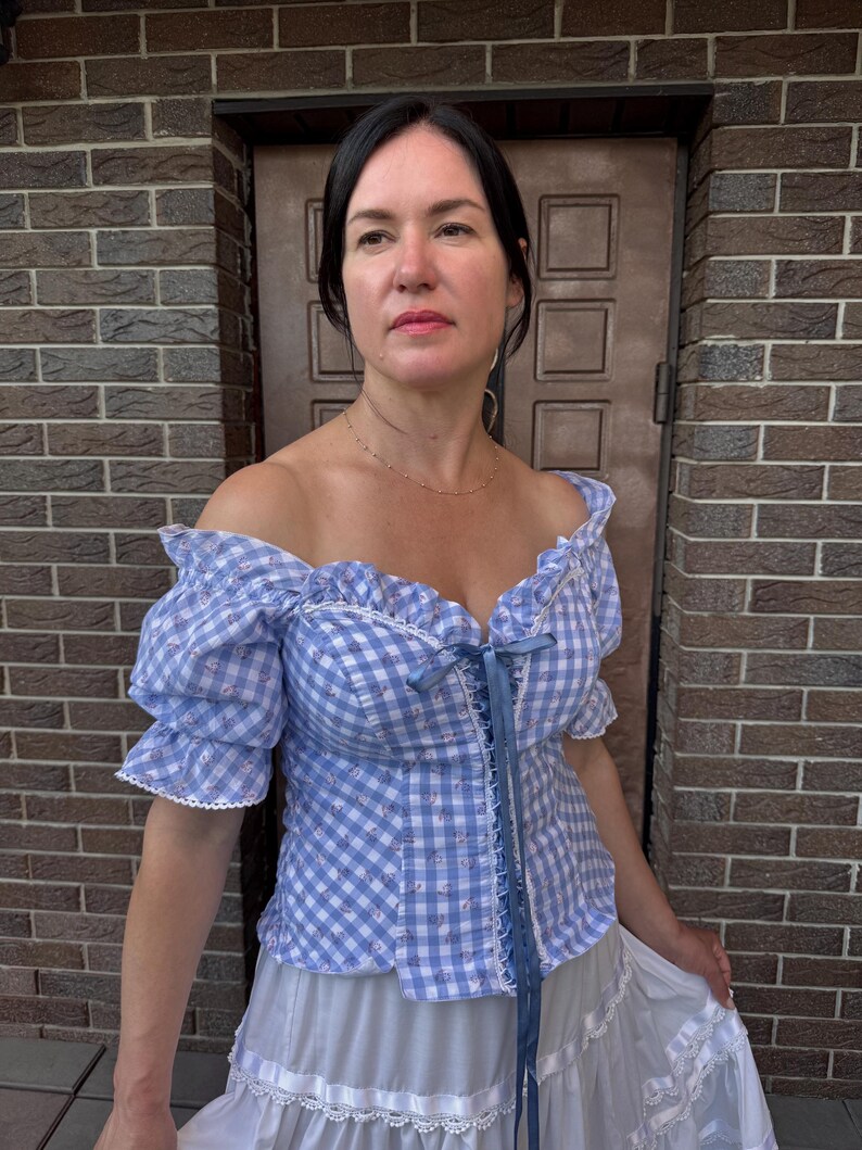 Puede incluir: Una blusa de hombros descubiertos en cuadros vichy azul claro y blanco, con mangas cortas abullonadas, escote coraz&oacute;n y un detalle de cordones con cinta azul en el centro. La tela tiene un estampado floral sutil y bordes de encaje blanco.