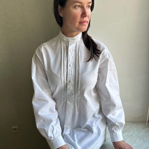 Puede incluir: Una camisa blanca de manga larga con cuello alto y cierre de botones en la parte delantera. La camisa tiene pliegues en la parte delantera y está hecha de una tela ligera.
