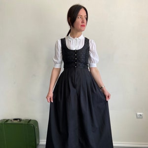 Puede incluir: Un corpi&ntilde;o negro con un sutil patr&oacute;n de puntos y una hilera de botones plateados, usado sobre una blusa blanca de manga corta con cuello redondo. El conjunto incluye una falda larga de color negro s&oacute;lido con cintura plisada hasta el suelo.