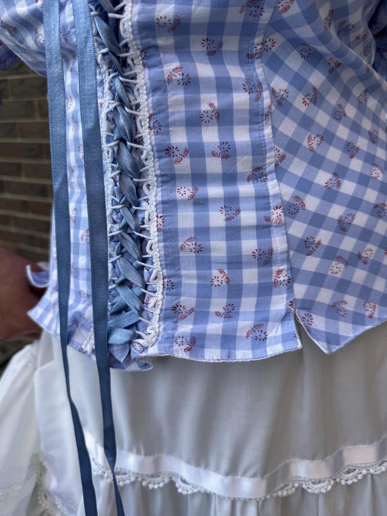 Puede incluir: Un top tipo cors&eacute; de cuadros vichy en azul claro y blanco con un peque&ntilde;o estampado floral rojo, cierre frontal con cordones de cinta de sat&eacute;n azul y ribetes decorativos blancos. La prenda se lleva sobre una falda blanca con bajo volanteado y encaje.
