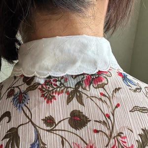 May include: A white collared blouse with a scalloped edge. The blouse is made of a sheer fabric with a floral print in shades of red, blue, and green.