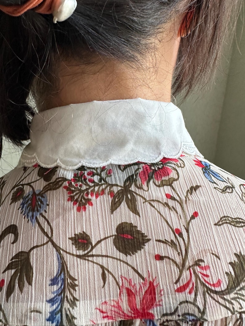 May include: A white collared blouse with a scalloped edge. The blouse is made of a sheer fabric with a floral print in shades of red, blue, and green.