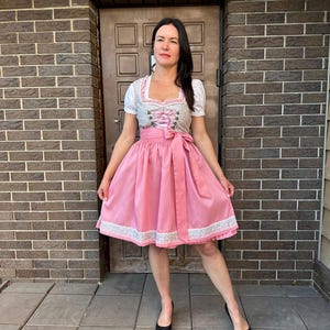 May include: A dirndl dress featuring a white blouse with short sleeves, a grey bodice with a floral pattern and pink lace-up detailing, and a solid pink skirt with a wide matching waist tie and a decorative white lace trim along the bottom hem.