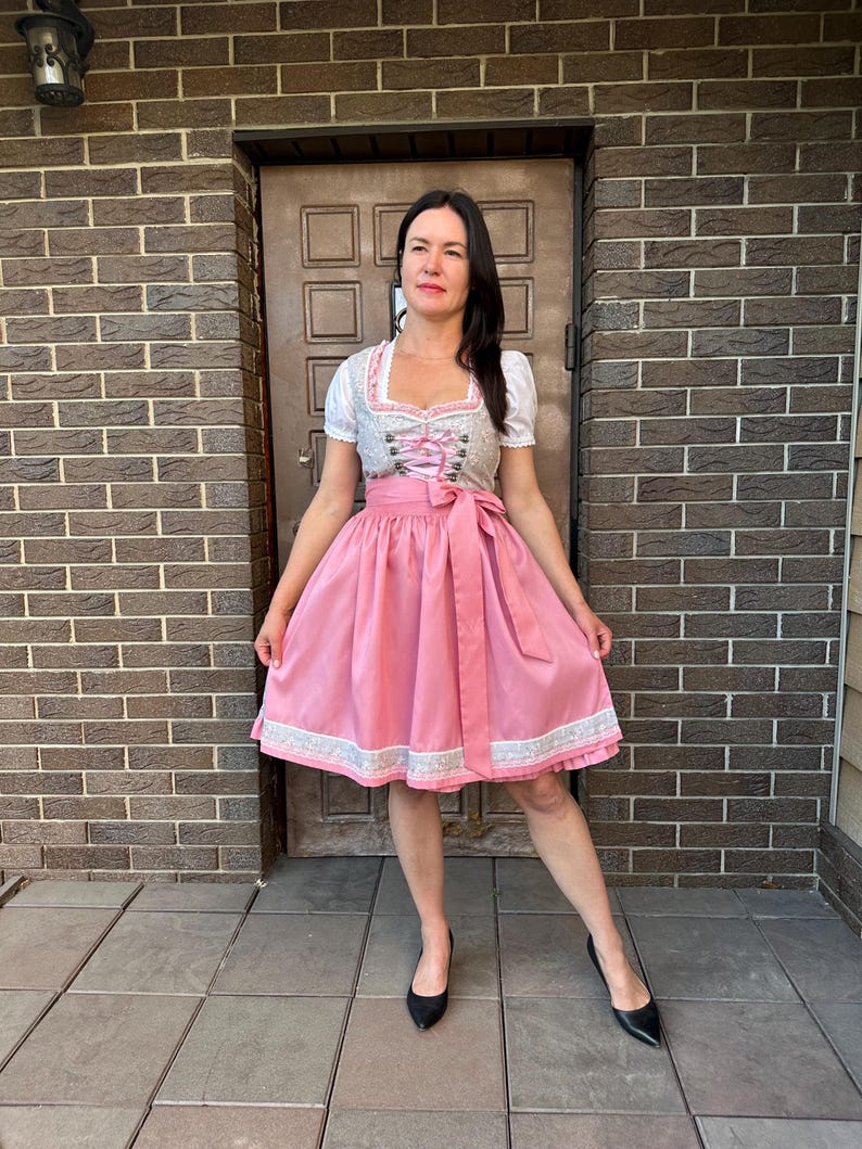 May include: A dirndl dress featuring a white blouse with short sleeves, a grey bodice with a floral pattern and pink lace-up detailing, and a solid pink skirt with a wide matching waist tie and a decorative white lace trim along the bottom hem.
