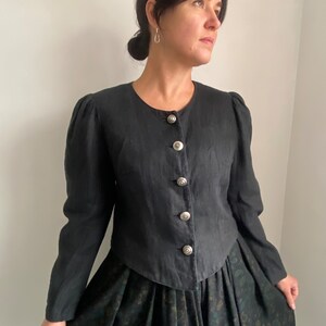 Puede incluir: Una chaqueta de lino negra con cuello redondo y botones plateados. La chaqueta tiene un corpi&ntilde;o ajustado y mangas largas. La modelo lleva la chaqueta sobre una falda floral verde oscuro.