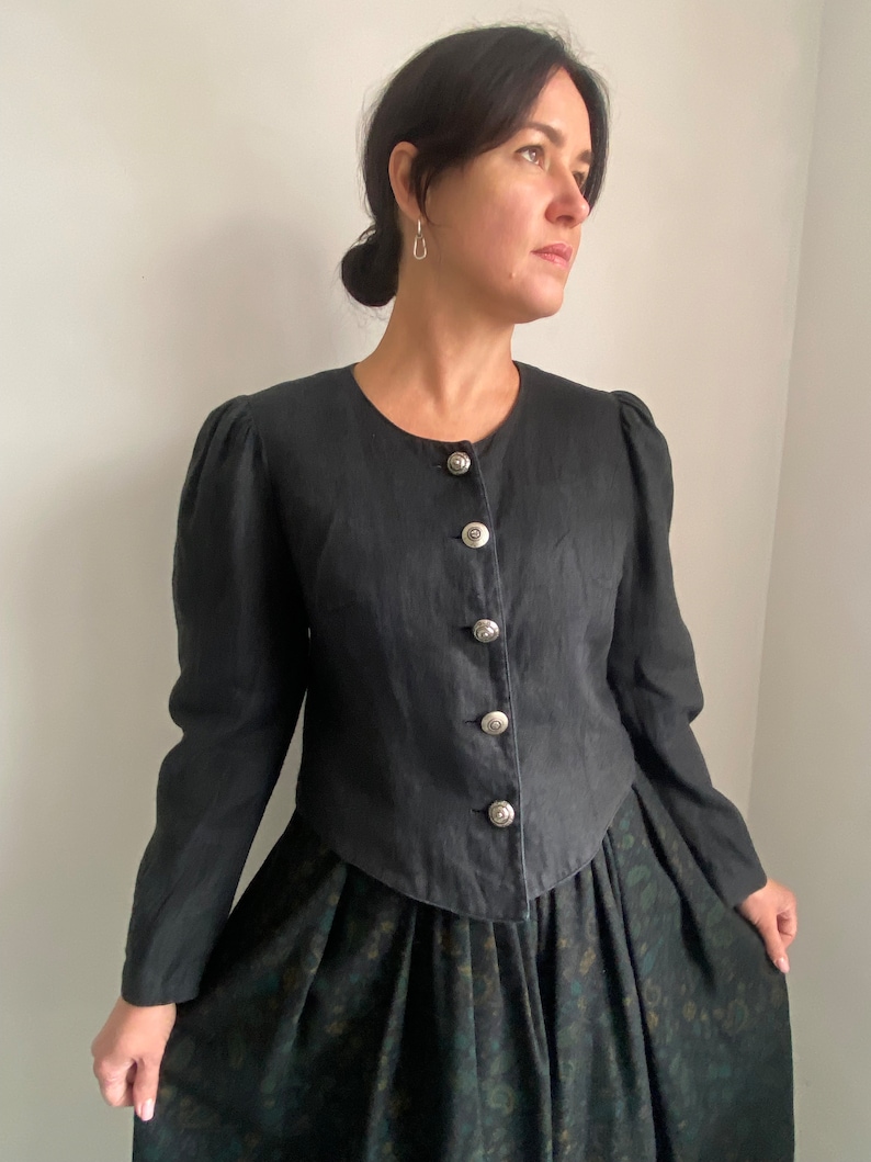 Puede incluir: Una chaqueta de lino negra con cuello redondo y botones plateados. La chaqueta tiene un corpi&ntilde;o ajustado y mangas largas. La modelo lleva la chaqueta sobre una falda floral verde oscuro.