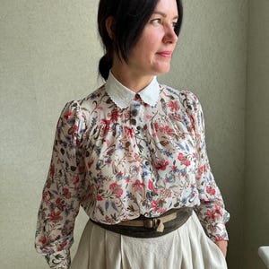 May include: A white floral print blouse with a white lace collar and long sleeves. The blouse is paired with a beige skirt and a brown leather belt.