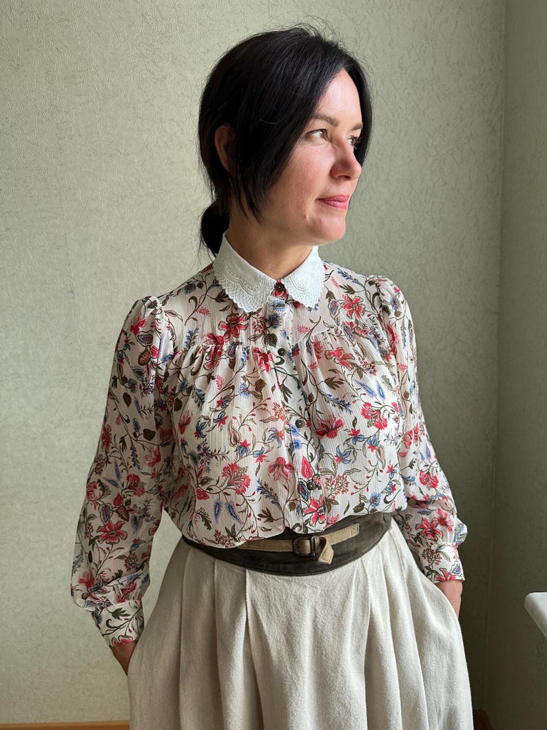 May include: A white floral print blouse with a white lace collar and long sleeves. The blouse is paired with a beige skirt and a brown leather belt.