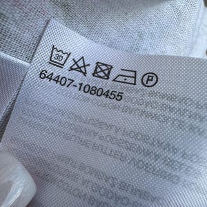 May include: A white fabric care label featuring a series of black laundry symbols including a 30-degree wash icon, a no-bleach triangle, a tumble dry square, an iron icon, and a dry clean circle, with the numerical code 64407-1080455 printed below.