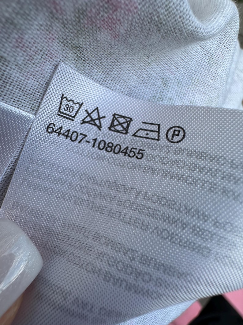 May include: A white fabric care label featuring a series of black laundry symbols including a 30-degree wash icon, a no-bleach triangle, a tumble dry square, an iron icon, and a dry clean circle, with the numerical code 64407-1080455 printed below.