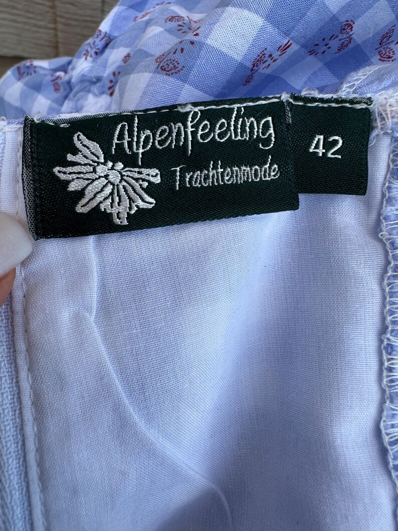 Puede incluir: Una prenda de tela de cuadros vichy azul y blanca con una etiqueta interior negra que presenta el texto blanco Alpenfeeling Trachtenmode, un gr&aacute;fico de flor edelweiss blanca y el n&uacute;mero 42. La tela tiene un sutil patr&oacute;n floral rojo.
