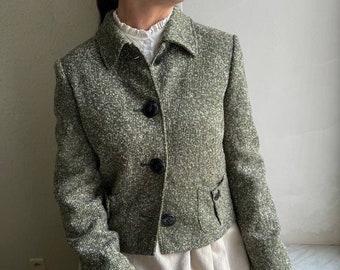Puede incluir: Una chaqueta de tweed verde recortada con botones negros y un pequeño bolsillo en la parte delantera.