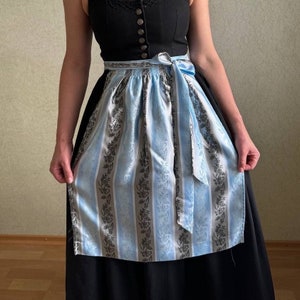 Könnte beinhalten: Ein schwarzes Kleid mit einer blau-weiß gestreiften Schürze. Die Schürze hat ein Blumenmuster und ein blaues Band, das in der Taille gebunden ist.