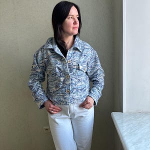 May include: A light blue jacket with a swirling pattern in shades of blue and white. The jacket has a button-up front, two chest pockets, and wooden buttons. The person is wearing white jeans.