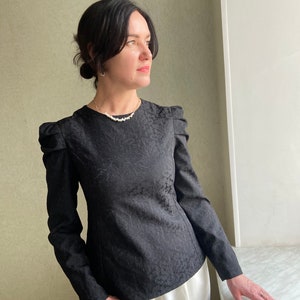 May include: A black long-sleeved blouse with a textured pattern and puffed shoulders. The blouse has a round neckline and is worn with white pants.