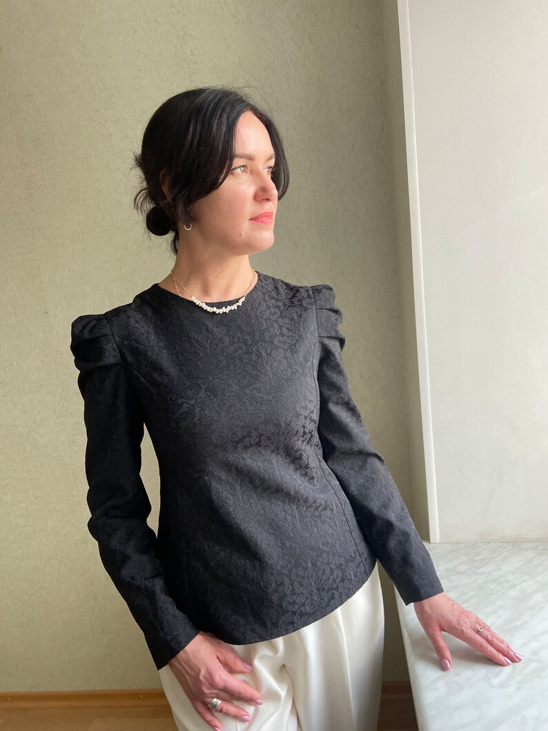 May include: A black long-sleeved blouse with a textured pattern and puffed shoulders. The blouse has a round neckline and is worn with white pants.