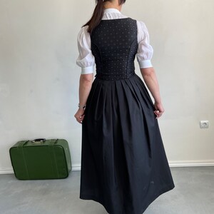 Puede incluir: Un corpi&ntilde;o negro con un sutil patr&oacute;n circular rojo combinado con una blusa blanca de manga corta con cuello alto y una falda negra larga y plisada. El conjunto se lleva con zapatos de tac&oacute;n color canela y una maleta vintage verde.