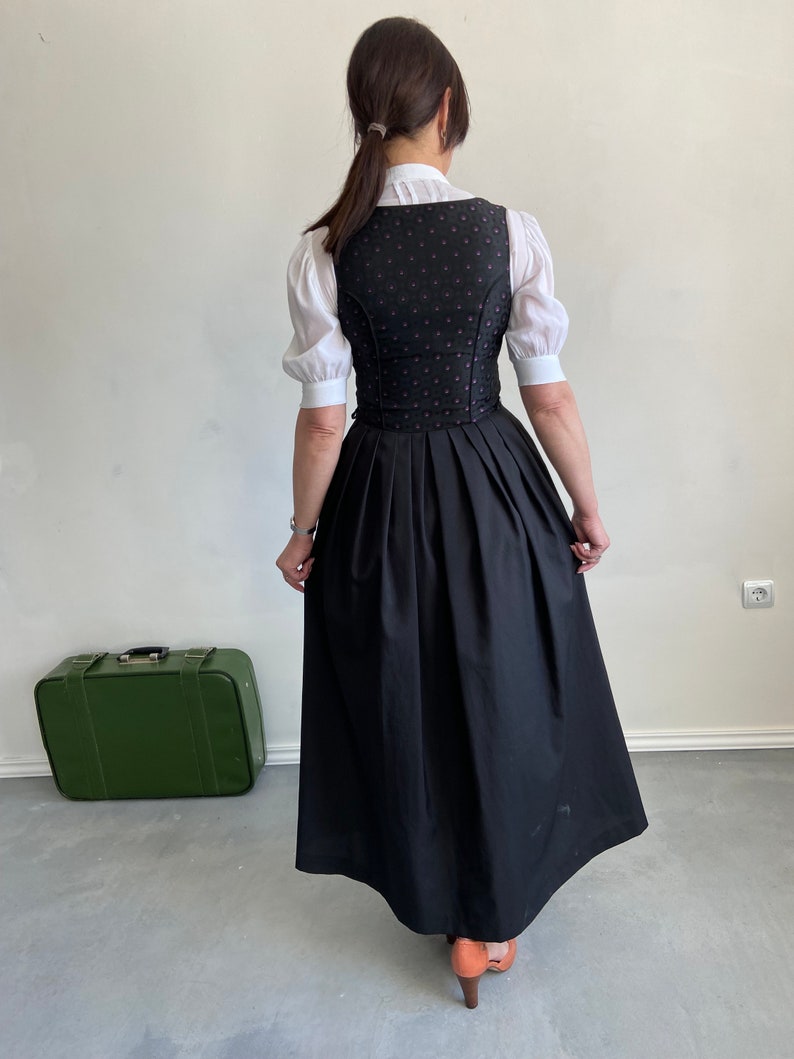 Puede incluir: Un corpi&ntilde;o negro con un sutil patr&oacute;n circular rojo combinado con una blusa blanca de manga corta con cuello alto y una falda negra larga y plisada. El conjunto se lleva con zapatos de tac&oacute;n color canela y una maleta vintage verde.