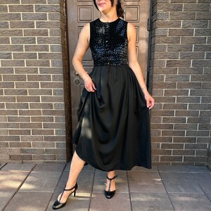 May include: A black sleeveless dress with a velvet bodice and a flowing skirt. The dress has a button closure at the front and a belt at the waist. The model is wearing black heels.