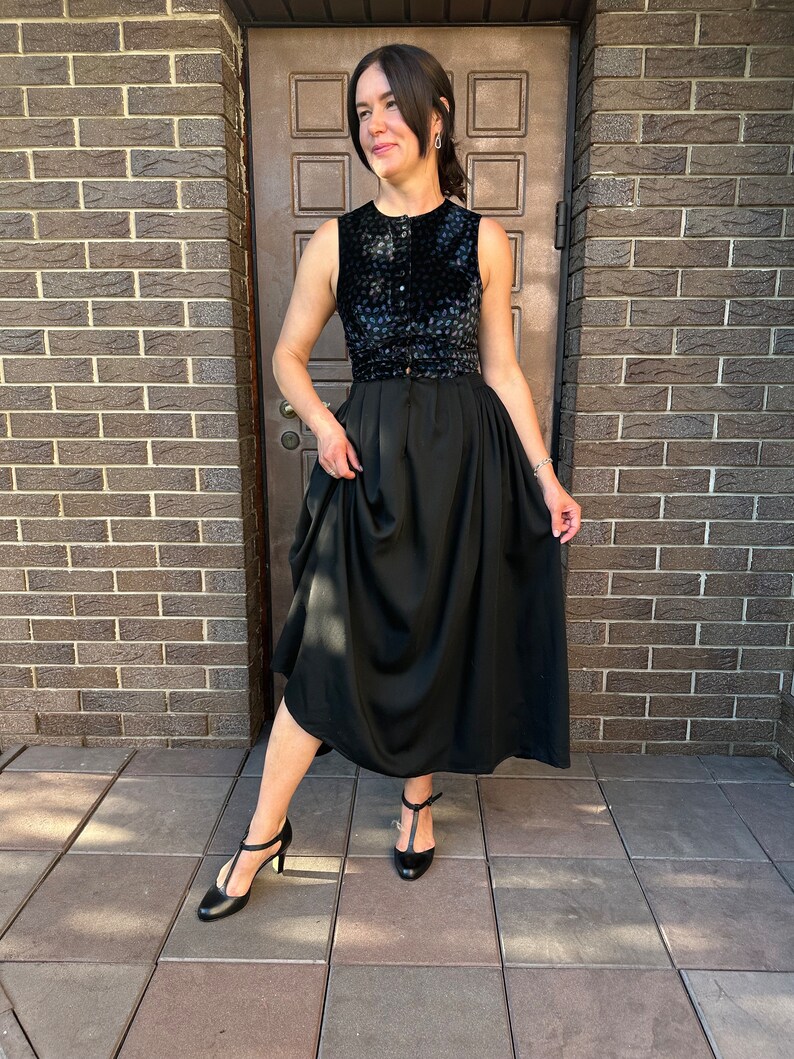 May include: A black sleeveless dress with a velvet bodice and a flowing skirt. The dress has a button closure at the front and a belt at the waist. The model is wearing black heels.
