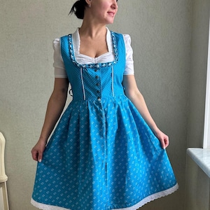 May include: A traditional blue dirndl dress with white floral patterns and a white lace trim. The dress features a square neckline, button-down front, and a white blouse underneath. The dress is a classic example of Bavarian folk attire.