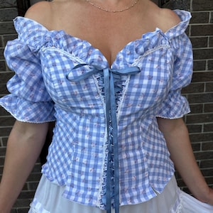 Puede incluir: Una blusa de hombros descubiertos con un patr&oacute;n de cuadros vichy en azul claro y blanco con peque&ntilde;os detalles florales. La prenda incluye mangas con volantes, ribetes de encaje blanco en el escote y las mangas, y un cord&oacute;n central de cinta azul.
