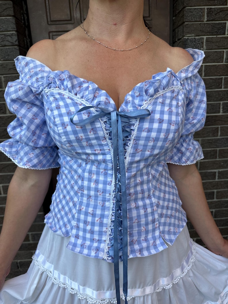 Puede incluir: Una blusa de hombros descubiertos con un patr&oacute;n de cuadros vichy en azul claro y blanco con peque&ntilde;os detalles florales. La prenda incluye mangas con volantes, ribetes de encaje blanco en el escote y las mangas, y un cord&oacute;n central de cinta azul.