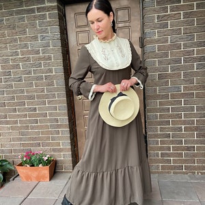 Puede incluir: Un vestido maxi marrón con un cuello de encaje blanco y un dobladillo con volantes. El vestido tiene mangas largas con puños de encaje. La modelo lleva botas negras y sostiene un sombrero de paja.