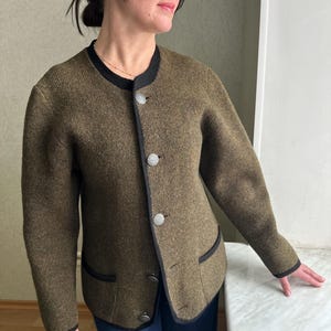 Könnte beinhalten: Olivgrüne Jacke mit schwarzem Besatz und silbernen Knöpfen. Die Jacke hat einen runden Ausschnitt und zwei Vordertaschen. Die Ärmel sind lang und der Stoff scheint eine Wollmischung zu sein. Ein klassisches, maßgeschneidertes Oberbekleidungsstück.