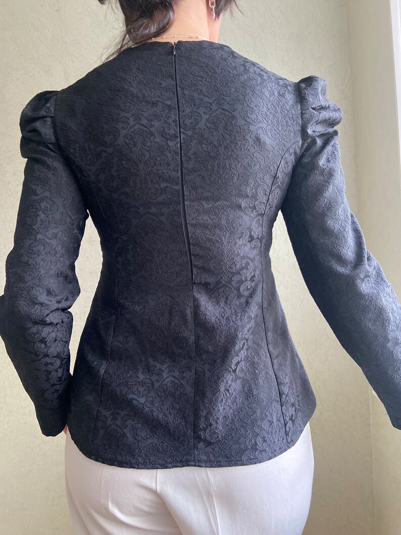 May include: Black long-sleeved blouse with a fitted silhouette and a subtle floral pattern. The blouse has a round neckline and a zipper closure in the back.