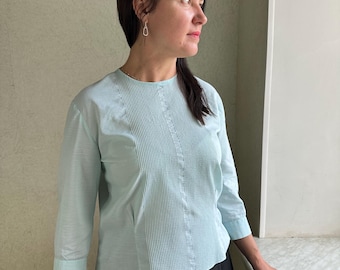 May include: A light blue, long-sleeved blouse with a round neckline and vertical pleats down the front. The blouse has a fitted silhouette and is worn with a black skirt.