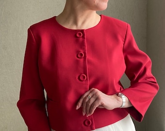 Puede incluir: Chaqueta roja con cuello redondo y mangas largas, abrochada con tres botones. Las mangas son acampanadas. La chaqueta se lleva con pantalones blancos. La chaqueta es de color liso. El diseño de la chaqueta es clásico y sencillo.