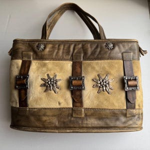 May include: A brown leather tote bag with a light brown suede panel and silver metal edelweiss accents. The bag has brown leather straps and buckles.