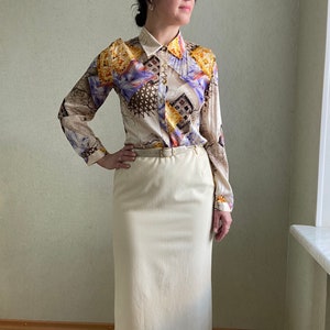 May include: A two-piece outfit featuring a long-sleeved button-down shirt and a knee-length skirt. The shirt has a cream-colored background with a colorful abstract pattern. The skirt is a solid cream color and is paired with a thin belt.