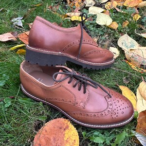 May include: Two brown leather Oxford shoes with decorative perforations and black laces, set on a bed of green grass and fallen autumn leaves. The shoes have a classic design with a sturdy sole.