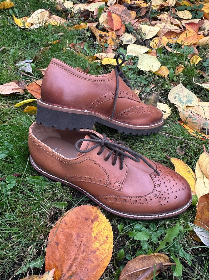 May include: Two brown leather Oxford shoes with decorative perforations and black laces, set on a bed of green grass and fallen autumn leaves. The shoes have a classic design with a sturdy sole.