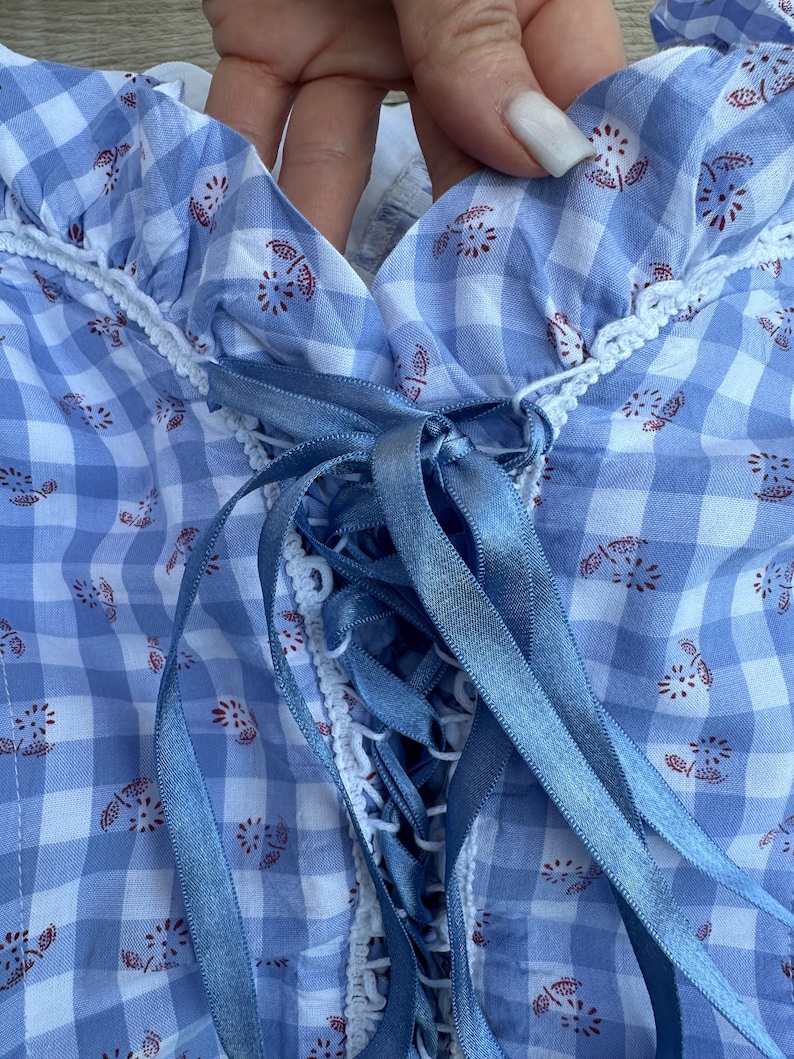 Puede incluir: Una blusa con estampado de cuadros vichy en azul claro y blanco con un peque&ntilde;o estampado floral rojo, escote con volantes y borde de ganchillo blanco, y un cierre central de cordones de cinta de sat&eacute;n azul atado en un lazo.