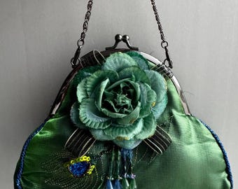 May include: A green satin evening bag with a decorative green flower and a peacock feather design. The bag has a silver chain handle and a blue beaded trim. The flower is accented with gold and blue details.