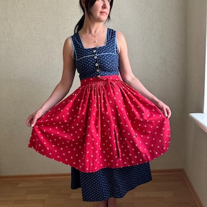 Puede incluir: Un vestido sin mangas con escote cuadrado. El corpiño es azul marino con lunares blancos y ribete blanco. La falda es roja con estampados florales blancos. El vestido tiene un lazo rojo a juego en la cintura. El vestido es hasta la rodilla.