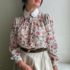 May include: A white blouse with a floral print and a white lace collar. The blouse has long sleeves and is cinched at the waist with a brown belt. The model is wearing a white skirt.