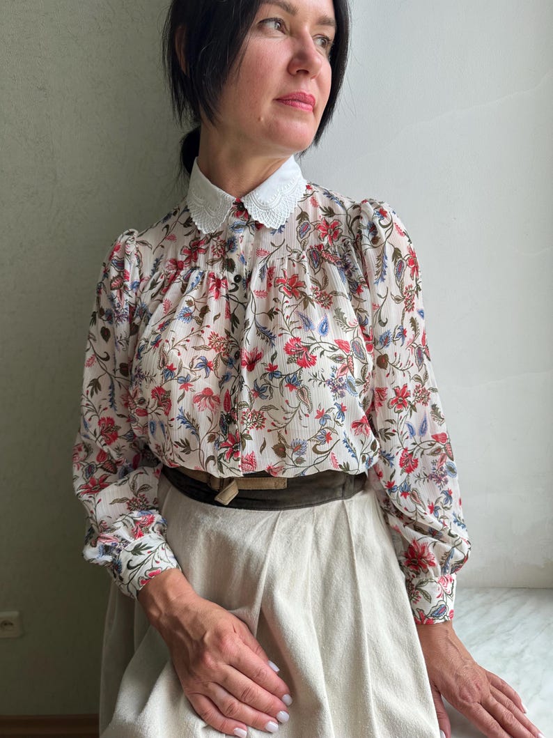 May include: A white blouse with a floral print and a white lace collar. The blouse has long sleeves and is cinched at the waist with a brown belt. The model is wearing a white skirt.